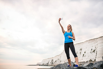 Young sporty woman runner with earphones and smartphone on the beach, taking selfie.