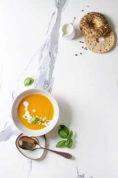 Bowl of vegetarian pumpkin carrot soup served with herbs, spoon, jug of cream, bagel bread, black pepper, salt over white marble texture background. Flat lay, space