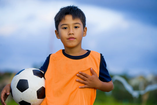 lifestyle portrait of handsome and happy young boy holding soccer ball playing football outdoors at green grass field smiling cheerful wearing training vest