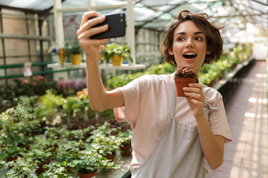 Woman gardener standing over flowers plants in greenhouse make selfie by mobile phone.