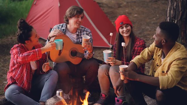 Joyful young people friends are clinking glasses with drinks sitting around fire in forest with warm marshmallow on sticks, smiling man is holding guitar.