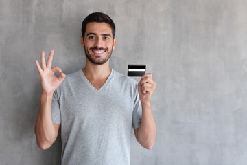 Closeup portrait of young handsome Caucasian guy pictured isolated on gray background, smiling, showing ok sign with his hand while holding debit or credit card in another one, feeling satisfied