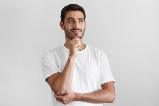 Horizontal shot of young European guy pictured isolated on grey background standing in blank T-shirt against wall, pressing fist to jaw as if dreaming of something, looking pensive and happy