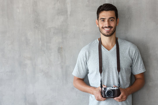 Horizontal Picture Of Handsome Caucasian Guy Pictured Isolated Against Grey Textured Wall In Grey T-shirt And Trousers With Camera Hanging On Strap On His Neck, Smiling As If Eager To Take Photos