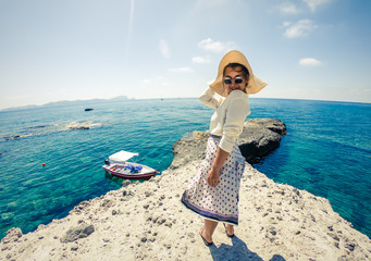 Young woman on rocky cliff in Palmarola island in front of the ocean on a sunny day. Elegant white dress with skirt and hat.