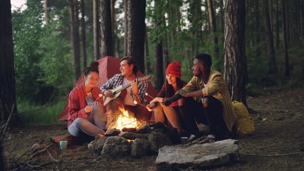 Tourist is playing the guitar sitting near campfire with friends singing and having fun, young people are holding sticks with marshmallow above flame. Food, music and fun concept.