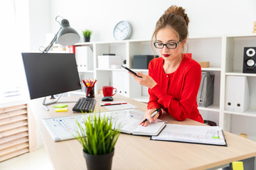 A young girl is standing at a table in the office, holding a black marker and a phone in her hand....