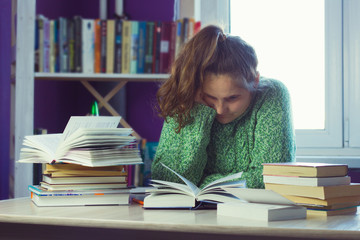 sad woman in a green sweater at a table in the middle of a stack of books and study materials, a blurred bookcase and a window for the background