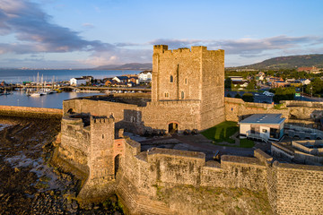 Medieval Norman Castle in Carrickfergus near Belfast in sunrise light. Aerial view with marina, yachts, parking, town and far view of Belfast in the background - obrazy, fototapety, plakaty