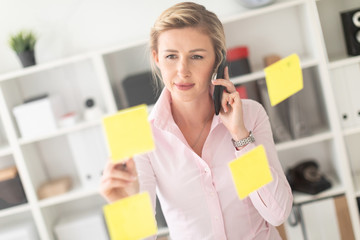 A young blonde girl is standing in the office next to a transparent board with stickers, holding a pencil in her hand and talking on the phone.