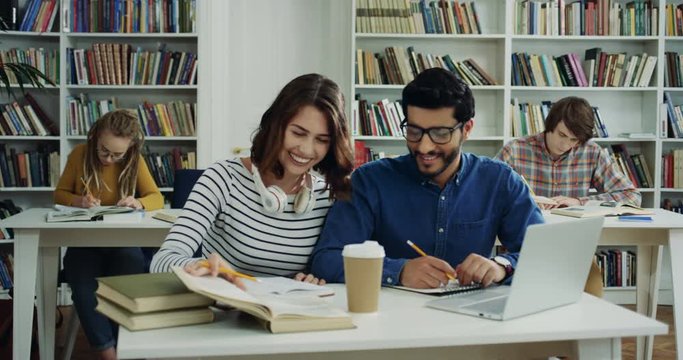 Couple of mixed races young man and woman studing together in the library and talking cheerfully.