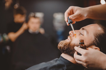 Barber applies beard oil with dropper for man in barbarshop.