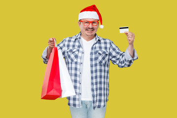 Happy modern casual style middle aged man in red new year cap, eyeglasses, standing and holding shopping gift bags and credit card with toothy smile, looking at camera. isolated on yellow background