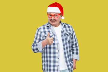 I like it! Optimistic casuald styled middle aged man in red santa cap, eyeglasses standing and showing thumbs up and toothy smiling, looking at camera. Studio shot, isolated on yellow background