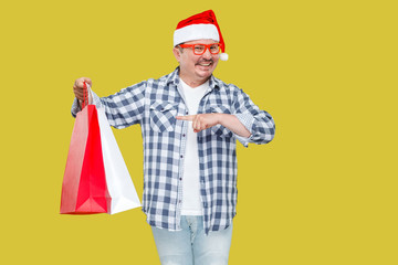 Casual styled funny middle aged man in red new year santa cap, eyeglasses standing and pointing finger to shopping bags and toothy smile, looking at camera. Studio shot, isolated on yellow background