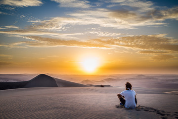 Fototapeta na wymiar Homme Assis de dos regardant un coucher de soleil à Hucachina au Pérou dans le désert