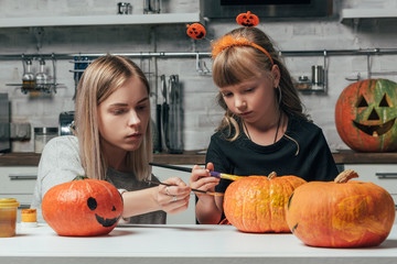 young woman and little sister painting pumpkins for halloween together in kitchen at home