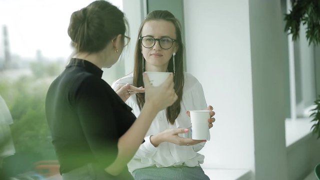 Two Business women near the window in the office drinking coffee at the end of the day and talk to hot gossip, commenting on the day.