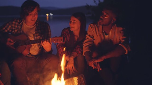 Happy friends are singing and playing the guitar getting warm around fire in forest near river on summer night. Millennials are wearing casual clothing.