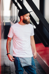 Fototapeta na wymiar Hipster handsome male model with beard wearing white blank t-shirt and a baseball cap with space for your logo