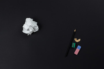 A flat lay photo of stationery set: pencil, rubber, sharpener and crumpled paper ball on the black background on the table in the office or school or university. Workspace of student or office worker.