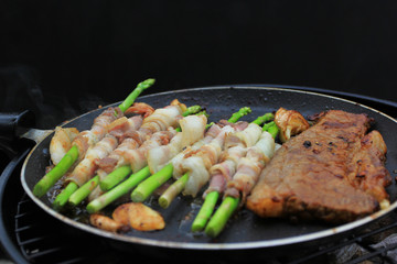 Raw bacon wrapped green asparagus and beef steak in frying pan on dark background