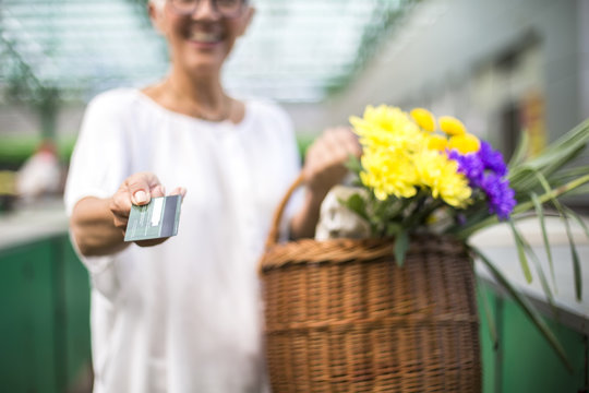 Senior woman holding credit card and  basket with bouquet of flowers on market