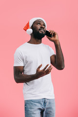 portrait of emotional african american man in santa claus hat talking on smartphone isolated on pink