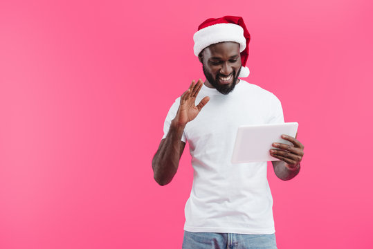 portrait of smiling african american man in santa claus hat with tablet gesturing isolated on pink - Powered by Adobe