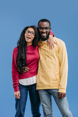 stylish happy african american couple in eyeglasses embracing each other isolated on blue background