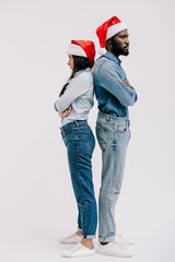 irritated african american couple in santa hats standing back to back with crossed arms isolated on white