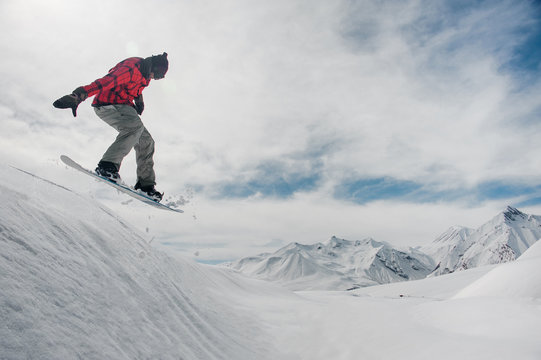 man is jumping on a snowboard against a snow-capped mountain peaks