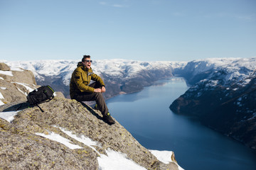 Top view of happy traveler man sitting on a rocky surface on a cliff and enjoying the scenic view of the fjord and mountains. Preikestolen, Pulpit rock, Lysefjord, Norway