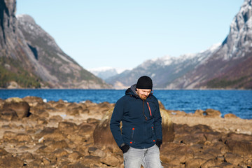 Young traveller man in winter jacket standing on the rocky shore of the fjord with beautiful view of the mountains with snow peaks. Fjord, Norway
