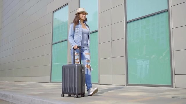 Confident, smiling woman walking on side walk with luggage wearing stylish denim clothes