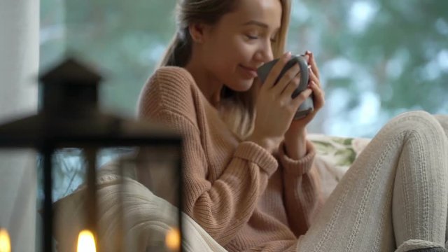 Cozy winter lifestyle. Happy young woman enjoy of cup of hot coffee sitting home by the big window with winter snow tree background
