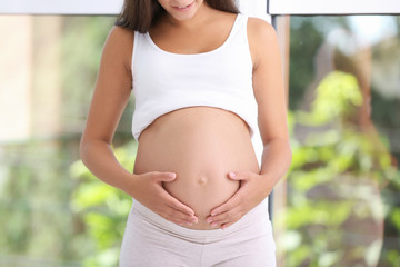 Pregnant woman standing near window at home, closeup