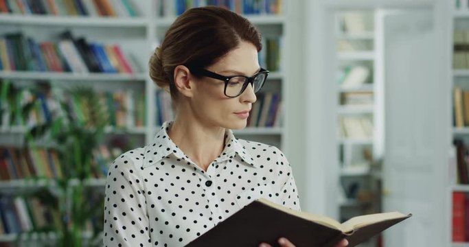Portrait of the Caucasian beautiful woman in glasses flipping pages and reading a book in the room full of the books on the shelves.