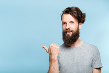 smiling young man pointing sideways with thumb as if showing smth. portrait of a bearded guy on blue background. copy space for advertisement.