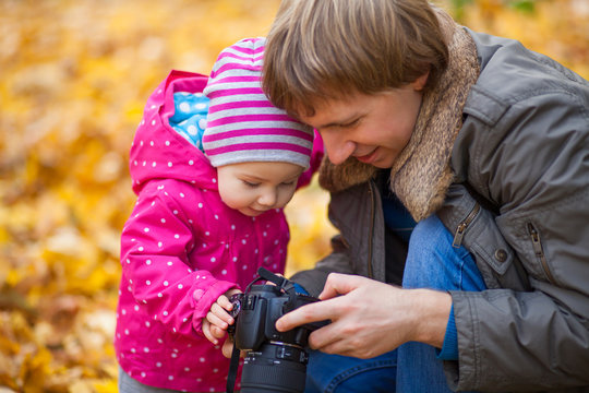 Little kid is playing with camera in autumn park. Daughter and father look at taking photos of autumn nature, landscapes. Baby girl is dressed in warm hat and jacket.