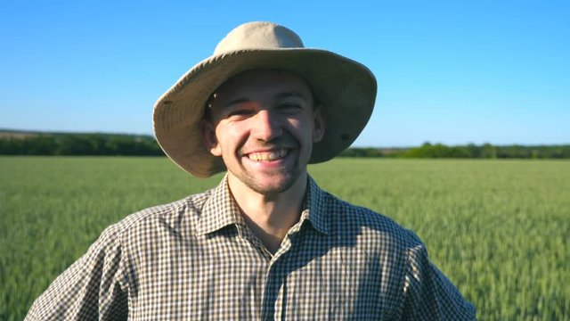 Close up of happy young farmer in hat looking and smiling into camera against the background of green wheat field. Portrait of handsome man in shirt standing in the meadow on sunny day