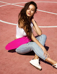 young sports happy sexy brunette girl with long hair in jeans and white top is sitting fashion and flirting with hand near her head with the pink mini skateboard on the pink sports court background