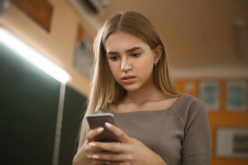 Nervous young student schoolgirl lady with long hair standing in empty classroom using mobile phone chatting.