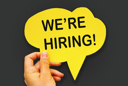 Hand holding yellow paper hand-cuted speech bubble with WE'RE HIRING concept, over gray background, isolated.