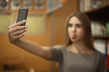 Funny young student schoolgirl lady with long hair standing in empty classroom take a selfie by mobile phone showing emotions tongue grimace. Focus on phone.