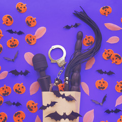 Sex toys are in a paper shopping bag. Nearby are scattered figures of pumpkins with funny faces, silhouettes of black bats and fallen leaves. Purple background. An image for a sex shop for Halloween.