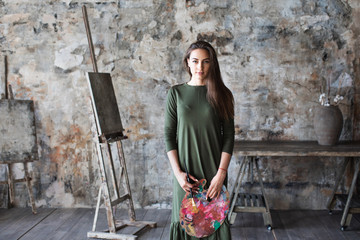 Young long-haired woman painter in a green dress with a brush and a palette in an art studio.
