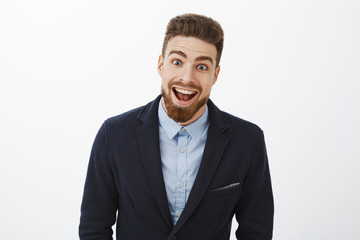 Waist-up shot of enthusiastic delighted and surprised charismatic young rich bachelor in stylish suit with beard and blue eyes smiling broadly from amazement and joy gazing impressed at camera