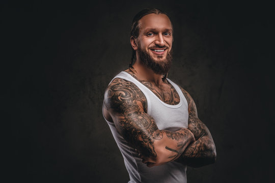 Smiling handsome bearded tattooed male in white shirt posing with crossed arms. Isolated on a dark textured background.