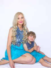 Mom and daughter in identical dresses are sitting on a white background, blondes. Beautiful family photo.